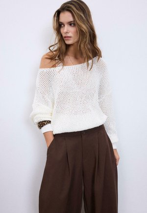 Donna che indossa un maglione bianco lavorato a maglia, ampio e scivolato sulle spalle, e pantaloni marroni larghi a vita alta, che guarda di lato con le mani in tasca.