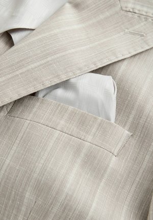 Giacca da completo beige a righe sottili con un fazzoletto da taschino grigio chiaro piegato ordinatamente nella tasca a petto.