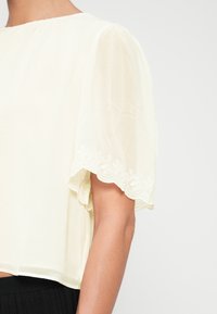 Haut du corps d'une personne portant un chemisier transparent blanc cassé avec des manches courtes bouffantes ornées de délicates broderies florales sur l'ourlet.