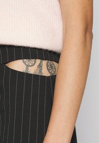 Sweater de malha rosa, curta na cintura, combinado com calças pretas de risca de giz de cintura alta com um recorte. Tatuagem visível por baixo do recorte.
