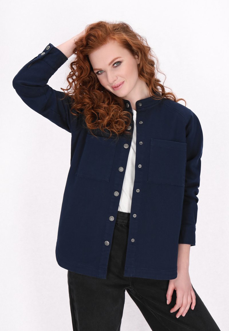 Camisa de botonadura navale con cuello alto, dos bolsillos frontales y dobladillo curvado. La tela parece suave y ligeramente estructurada.