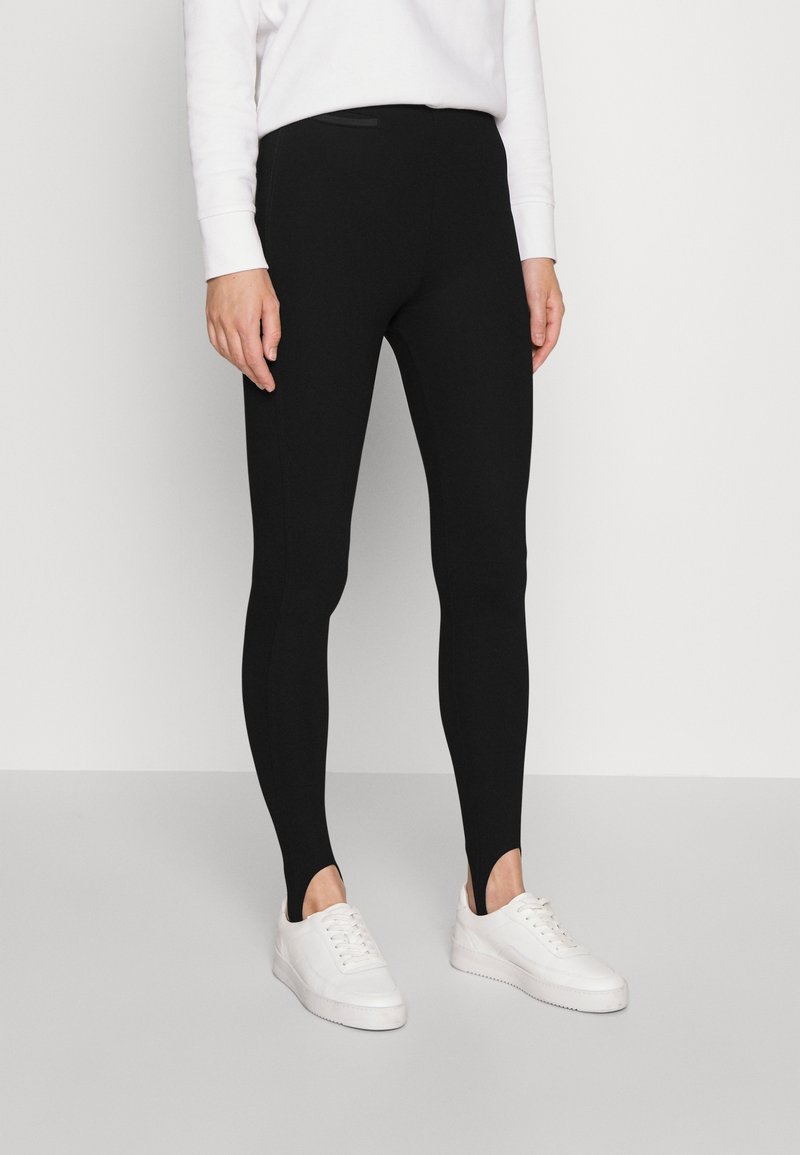 Personne portant des leggings noirs avec étriers et des baskets blanches, debout devant un fond clair uni.
