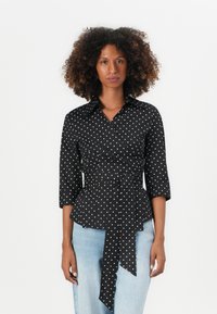 Camicia avvolgente nera a pois con maniche a tre quarti, vita aderente e dettaglio in vita annodato. Il tessuto ha una consistenza liscia.
