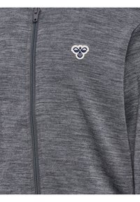 Grå lynlåstrøje i tekstureret stof med en lille broderet hvid og navy abstrakt bi-logo på venstre bryst.