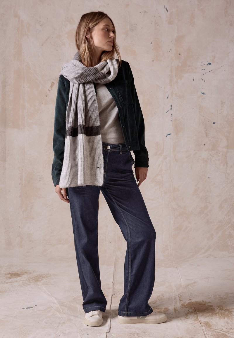 Una bufanda de punto a rayas grises y blancas cuelga sobre una chaqueta de corduroy verde, combinada con una blusa gris y unos jeans de pierna ancha azul oscuro. Los zapatos son color beige.