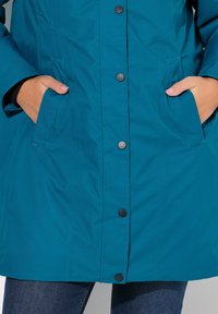 Teal vattentät parka med knappstängning, sidofickor och rak silhuett i texturerat tyg för värme.