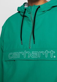 Grøn pullover jakke med hætte, med en lynlås foran, sorte snore og en fremtrædende hvid logo tværs over brystet.