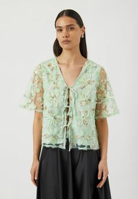 Lys grønn blomstret blonder topp med korte ermer, med blomstermønster og snøring i front. Gjennomsiktig tekstur med frynsete kanter.