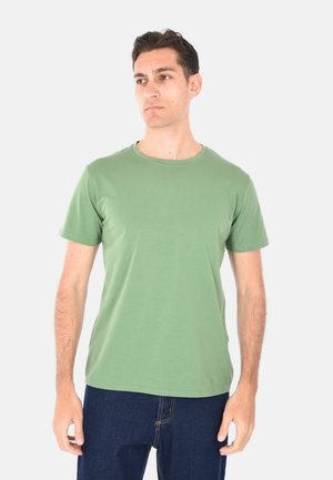 Uomo che indossa una t-shirt verde chiaro a maniche corte tinta unita e jeans blu scuro, in piedi e guarda leggermente a sinistra su sfondo bianco.