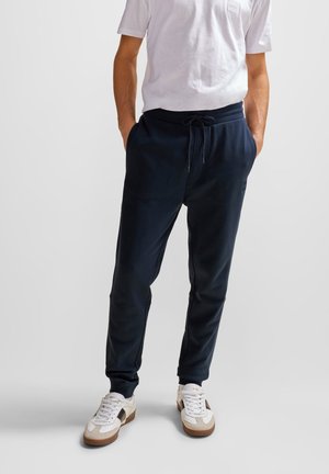 Mann trägt marineblaue Jogginghosen mit Kordelzug am Bund, weiße Turnschuhe und weißes kurzärmeliges T-Shirt, steht mit den Händen in den Taschen.