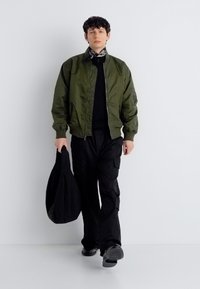 Άνδρας που φοράει πράσινο μπουφάν bomber, μαύρο cargo παντελόνι, μαύρα παπούτσια, κασκόλ με σχέδια γύρω από το λαιμό, κρατώντας μεγάλη μαύρη τσάντα, περπατάει προς τα εμπρός με φόντο λευκό.