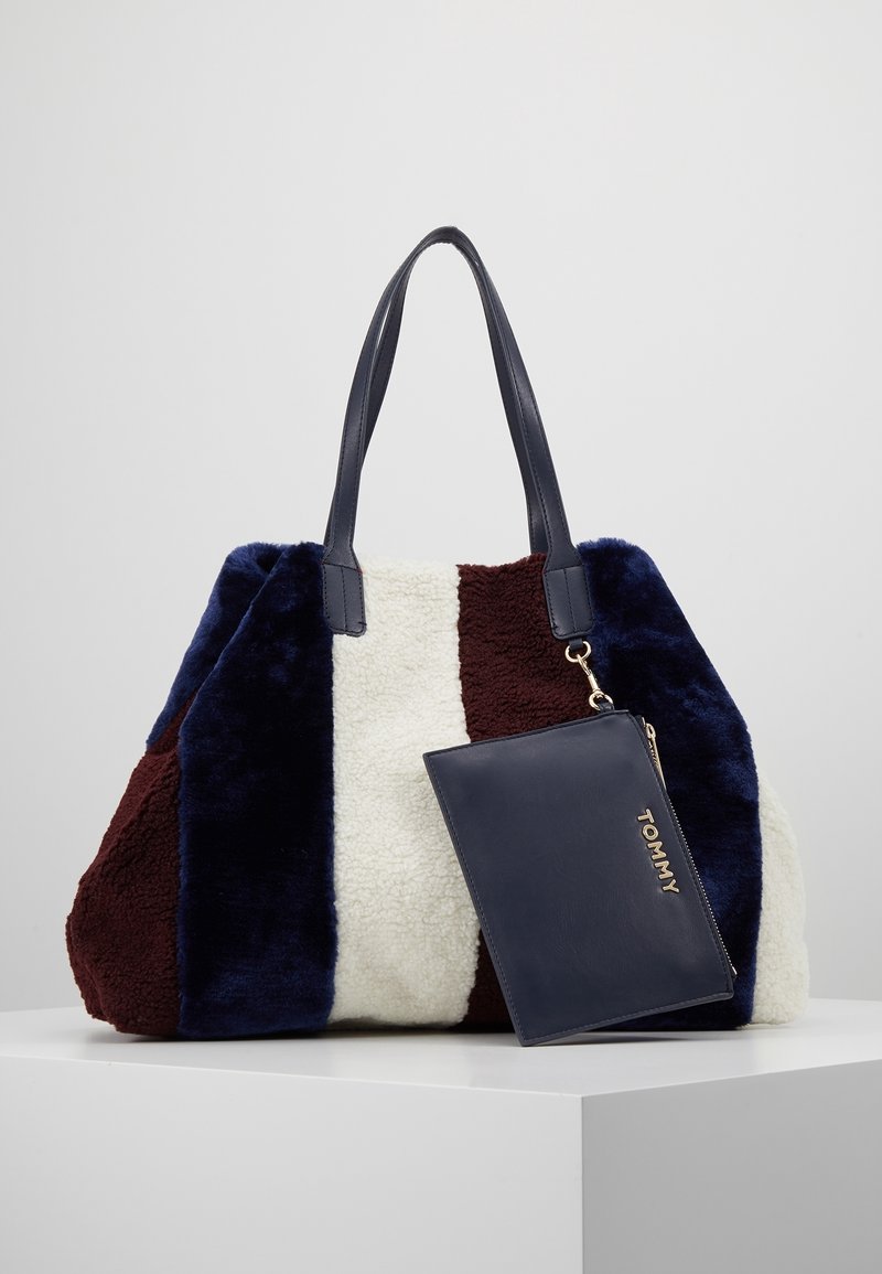 Sac cabas avec des rayures verticales en fausse fourrure bleu marine, blanche et bordeaux, poignées noires et une pochette noire étiquetée "TOMMY" attachée.