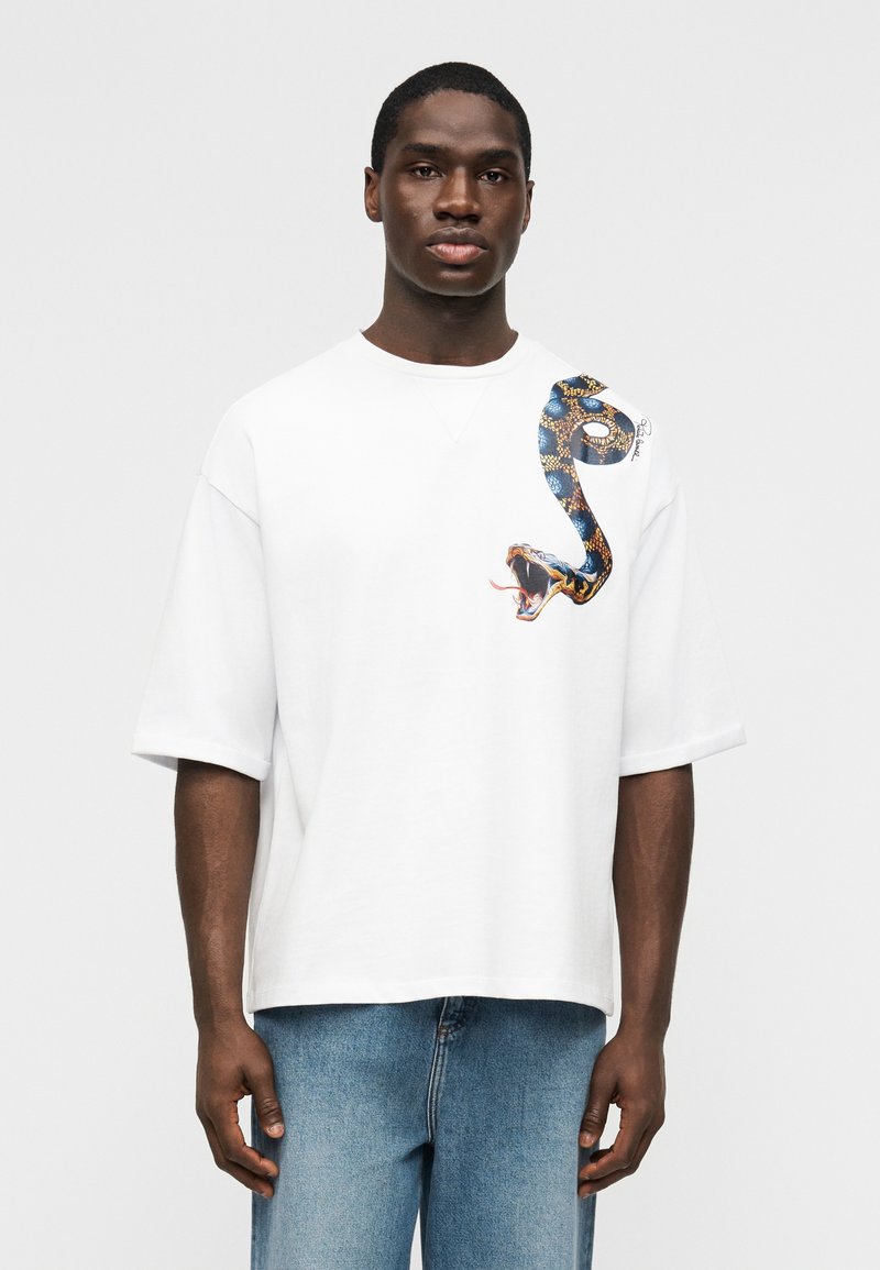 Άνδρας που φοράει ένα λευκό φαρδύ t-shirt με μεγάλο πολύχρωμο γραφικό φιδιού στον αριστερό ώμο, σε συνδυασμό με μπλε τζιν.