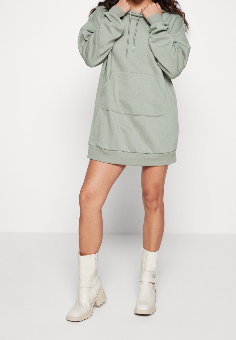 Femme portant une robe sweat à capuche oversize vert clair et des bottes blanches mi-mollet, debout devant un fond clair uni.