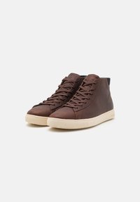 Brown leather high-top shoes with a textured finish, featuring a light-colored rubber sole and lace-up design. Minimalistic branding on side.
