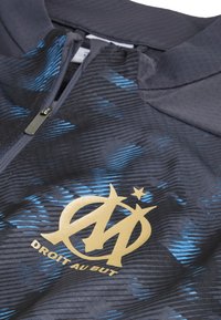 Zwarte jas met blauw patroon; heeft een ritssluiting en een gouden logo op de borst met de tekst "DROIT AU BUT" met een steraccent.