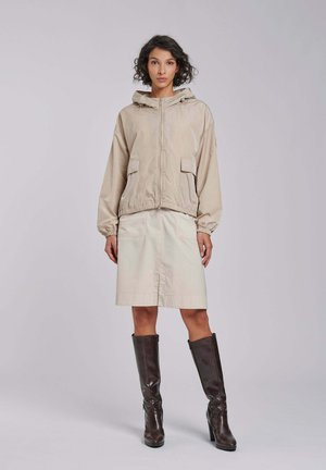 Kobieta o krótkich kręconych włosach, ubrana w beżową kurtkę z kapturem, kremową spódnicę do kolan oraz ciemnobrązowe kozaki na wysokim obcasie sięgające do kolan, stojąca na jednolitym tle.