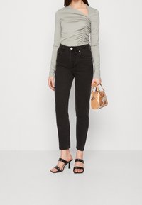 Maglietta grigia chiara a maniche lunghe con dettagli arricciati, abbinata a jeans neri a vita alta. La modella tiene in mano una piccola borsa color beige e indossa sandali neri con tacco.