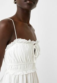 Mörkhudad person i en vit ärmlös klänning med tunna remmar, volanger och en knytning vid bröstet mot en enfärgad bakgrund.