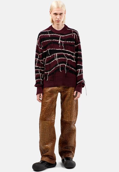 Maglione bordeaux con strisce bianche e nere, caratterizzato da dettagli sfilacciati; pantaloni marroni con texture effetto coccodrillo; scarpe nere slip-on.