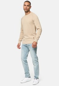 Suéter de punto beige de cuello redondo con puños y dobladillo acanalados, combinado con jeans azul claro y zapatillas blancas. Detalle notable del logotipo marrón en el suéter.
