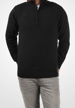 Uomo con maglione nero a trama con mezza cerniera e pantaloni grigio chiaro, mani dietro la schiena, su sfondo bianco uniforme.