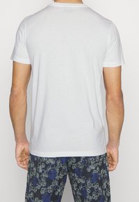 Biała bawełniana koszulka z krótkim rękawem, z okrągłym dekoltem. W połączeniu z ciemnoniebieskimi spodniami w wzory, ukazującymi kwiatowe i abstrakcyjne motywy.