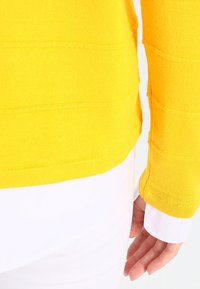 Maglione giallo a maglia con righe orizzontali texture, dotato di scollatura e polsini a coste. Indossato sopra una camicia bianca.