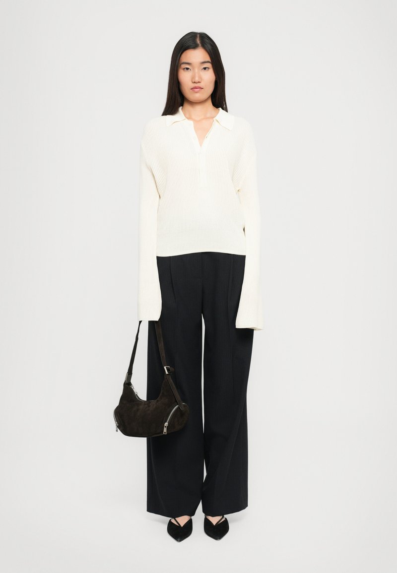 Maglione a coste bianche con scollatura a collo, abbinato a pantaloni larghi scuri. Una borsa in suede scuro e scarpe nere a punta completano il look.