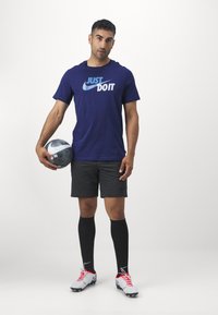 Navy T-shirt with blue "Just Do It" logo, black shorts, black knee-high socks, and white cleats with red accents, holding a black soccer ball.