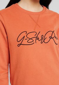 Sudadera naranja con logotipo de marca bordado en negro. Cuenta con cuello redondo, mangas largas y una textura suave. Tiene un sutil detalle en las costuras.