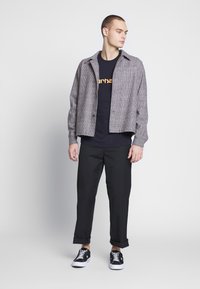 Chaqueta de patrón de espiga en gris y negro, combinada con una camiseta negra que presenta un texto gráfico en naranja, y pantalones negros a medida. Zapatillas blancas.