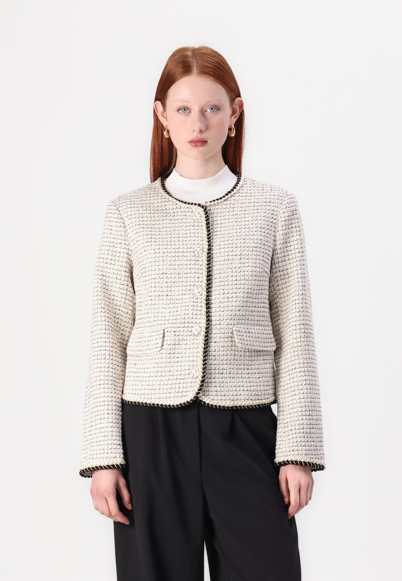 Giacca in tweed intrecciato di colore crema con accenti neri, scollatura tonda, tasche frontali e cuciture decorative lungo i bordi.