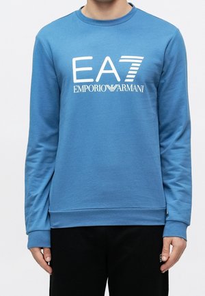 Blauwe sweatshirt met lange mouwen met wit "EA7 Emporio Armani" logo, gedragen door een persoon met zwarte broek tegen een effen witte achtergrond.