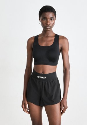Slank person med kort krøllet hår iført svart sports-bh og svarte Calvin Klein-shorts, stående mot en enkel lys bakgrunn.