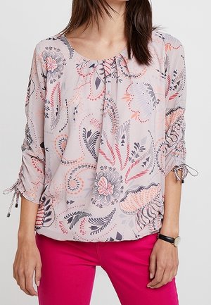 Blusa rosa chiaro con motivi floreali e geometrici, scollatura arricciata e maniche regolabili. Il tessuto sembra leggero e morbido.