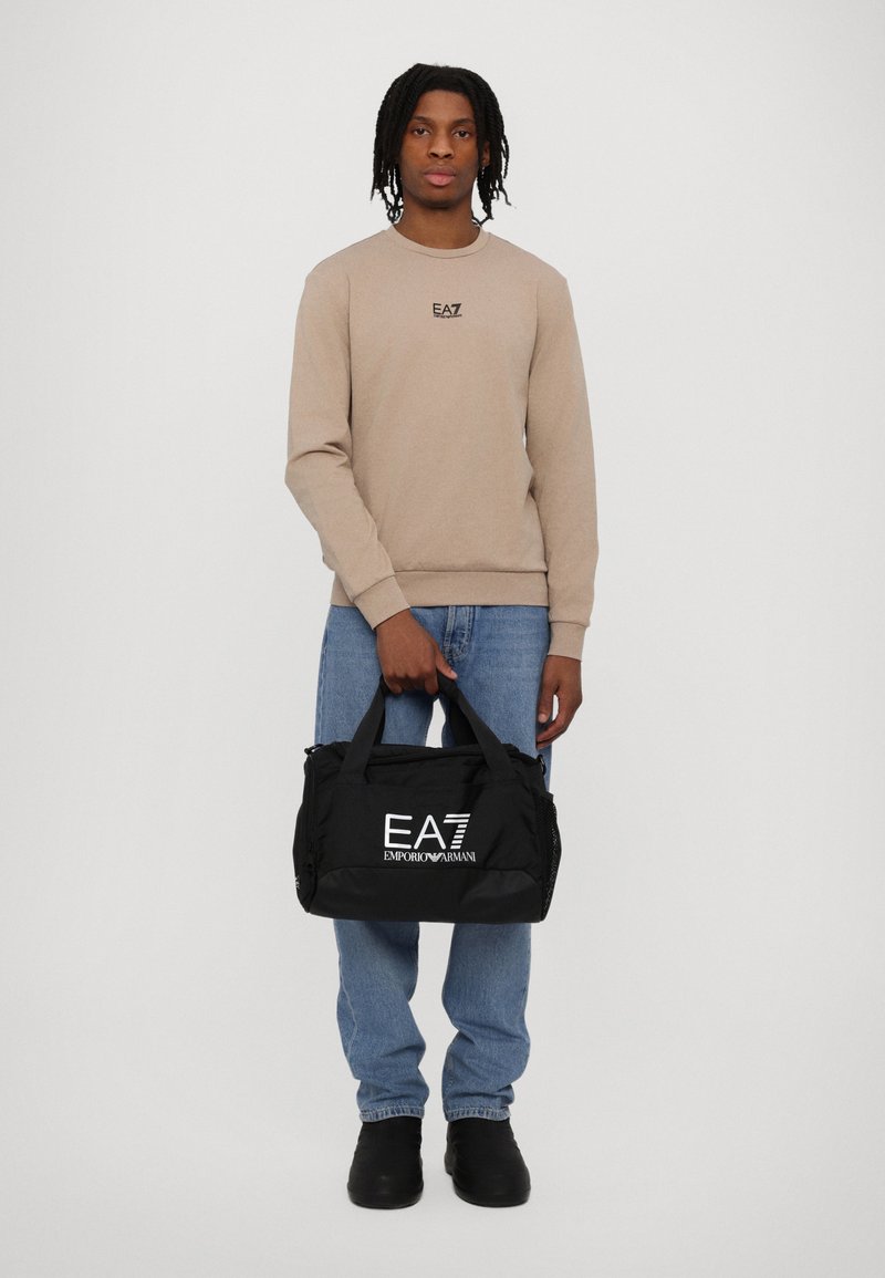 Svart duffelbag med hvitt "EA7 Emporio Armani"-logo, laget av slitesterkt materiale, holdt av en person iført beige sweatshirt og blå jeans.