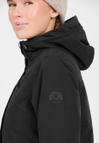 Svart huva jacka med en strukturerad yta, med en cirkulär logotyp på vänster ärm. Modell bär en ljusbeige mössa.