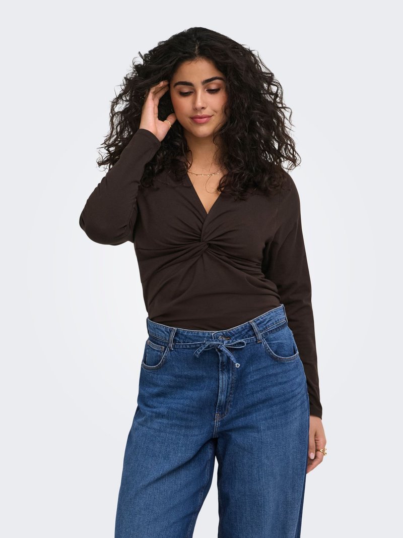 Femme aux cheveux bouclés portant un haut noir à manches longues avec un devant torsadé et un jean bleu taille haute sur un fond clair.