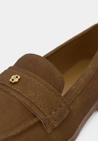 Mocasini maro din piele întoarsă, cu vârf rotund, cu un design cusut și un accent cu logo auriu în față. Căptușeală interioară netedă și talpă.