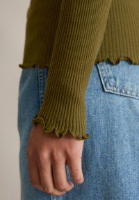 Haut côtelé vert olive avec ourlet froncé, assorti à un denim bleu clair. Gros plan sur la manche et la main, mettant en valeur la texture et l'ajustement.