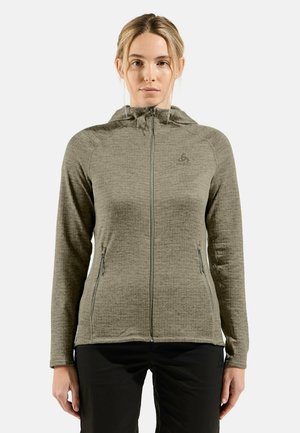 Woman wearing a light olive green zip-up hoodie with two front zippered pockets and black pants, facing forward against a white background.