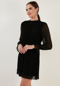 Mustapleated mekko, jossa on korkea kaula-aukko, läpinäkyvät pitkät hihat ja rypytetty vyötärö. Pehmeä, kevyt kangas, jolla on virtaava siluetti.