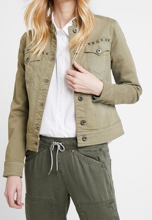 Lyshåret person iført en olivengrøn jakke med knapper over en hvid skjorte og matchende olivengrønne bukser med snøre i taljen.