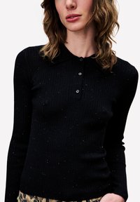 Vrouw in een zwart geribbeld poloshirt met lange mouwen met kleine multikleurige spikkels en een broek met luipaardprint tegen een witte achtergrond.