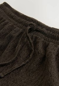 Gestructureerde donkerbruine stof van de broek, voorzien van een trekkoord met een knoop bij de tailleband, gelegd op een lichte achtergrond.
