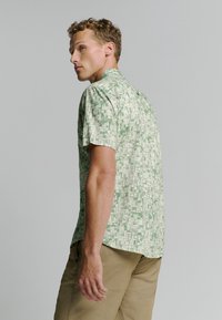 Korte mouwen shirt in lichtgroene stof met een geometrisch patroon. Heeft een casual kraag en een relaxed fit. Gecombineerd met lichtbeige broeken.