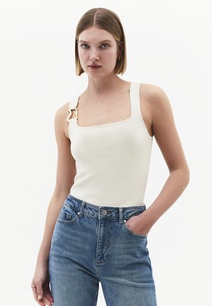 Jeune femme aux cheveux courts portant un top blanc sans manches avec une boucle dorée sur une bretelle et un jean bleu taille haute, une main dans la poche.
