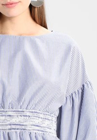 Blå och vitrandig blus med vid halsringning, puffärmar och smockad midjedetalj, tillverkad av lättviktig tyg.