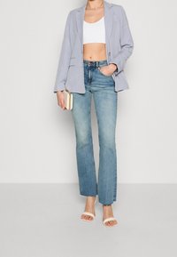 Jasnoszara marynarka na białym krótkim topie, zestawiona z jasnoniebieskimi dzwonami i białymi sandałami na obcasie. W jednej ręce elegancka kopertówka.